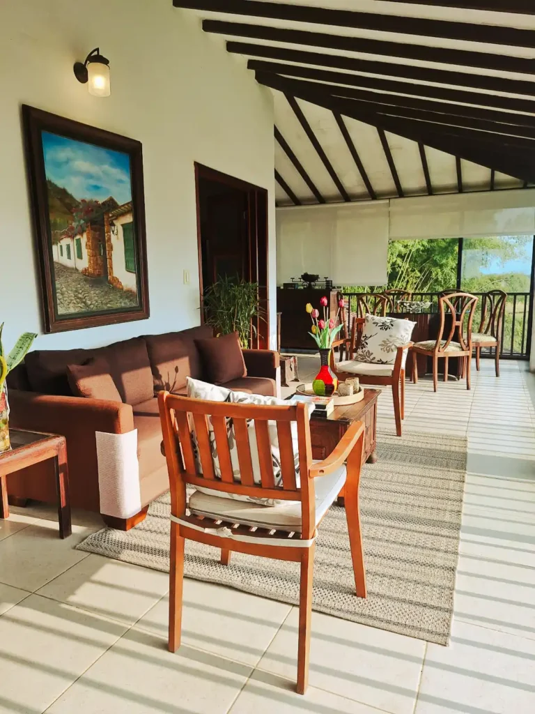 Zona común luminosa de hotel o finca en Alcalá, Valle del Cauca, con asientos cómodos y acceso a terraza con vista verde.